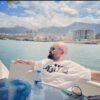 Man in white shirt and sunglasses sitting on boat, overlooking coastal city and mountains, blue sky.