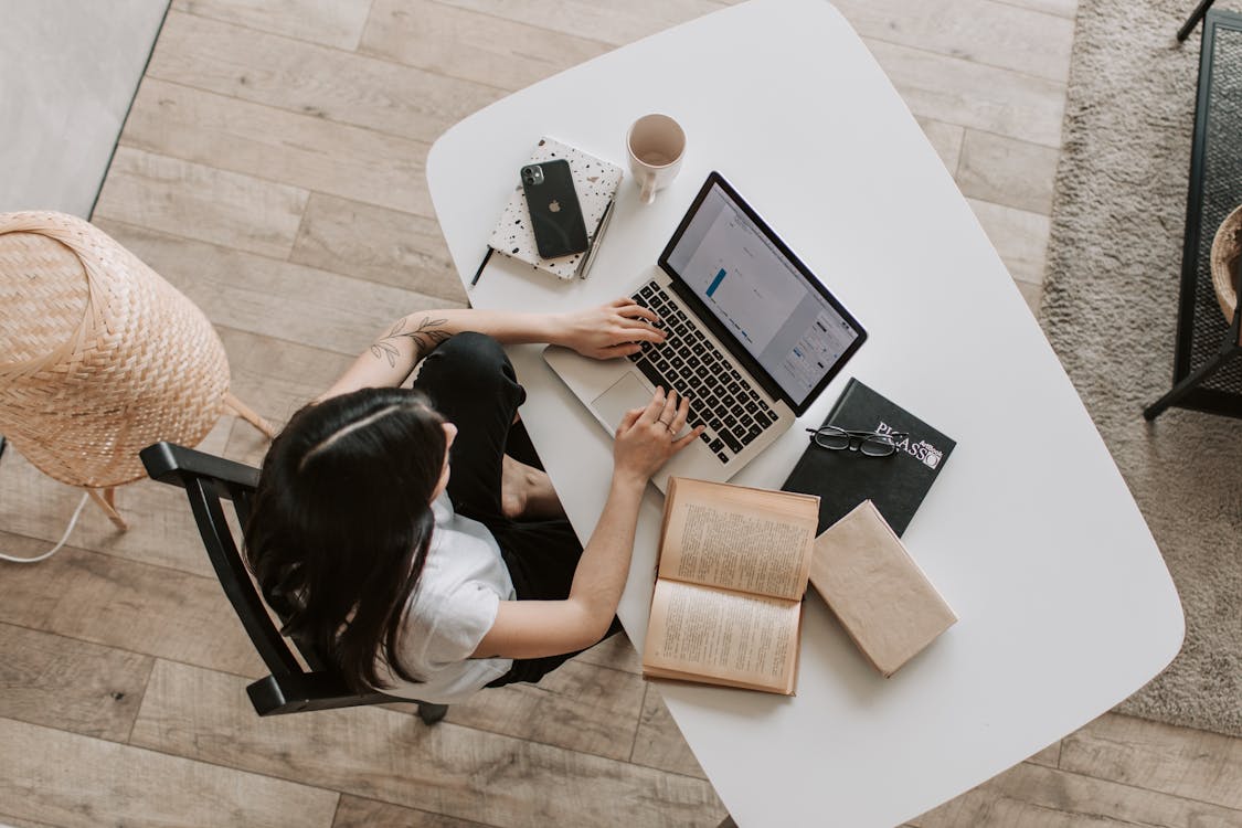 Person using laptop on white table with paper books, ceramic mug, smartphone, and black chair on wooden floor