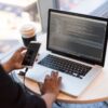 Laptop with metal body displaying code, person holding white smartphone, wooden round table with coffee cup and glasses