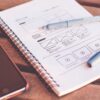 spiral notebook with paper wireframe sketches, plastic blue pen, and black smartphone on wooden surface