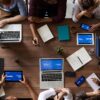 Wooden table with laptops, tablets, smartphones, notebooks, coffee mugs, and people working together