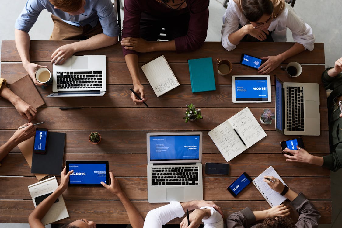 Wooden table with laptops, tablets, smartphones, notebooks, coffee mugs, and people working together