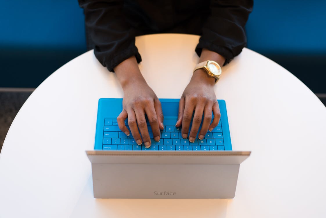 Person typing on blue plastic keyboard attached to silver laptop on white round table