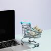 miniature metal shopping cart with blue handle holding rolled US dollar bills next to silver laptop