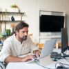 man in wheelchair using silver laptop on white desk in modern office with black monitor