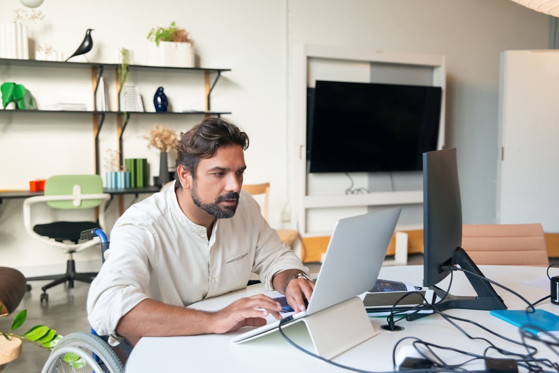 man in wheelchair using silver laptop on white desk in modern office with black monitor