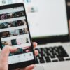 Hand holding black smartphone with photo gallery app open, in front of silver laptop keyboard