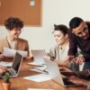 group of people using laptops and tablet around wooden table in office with corkboard