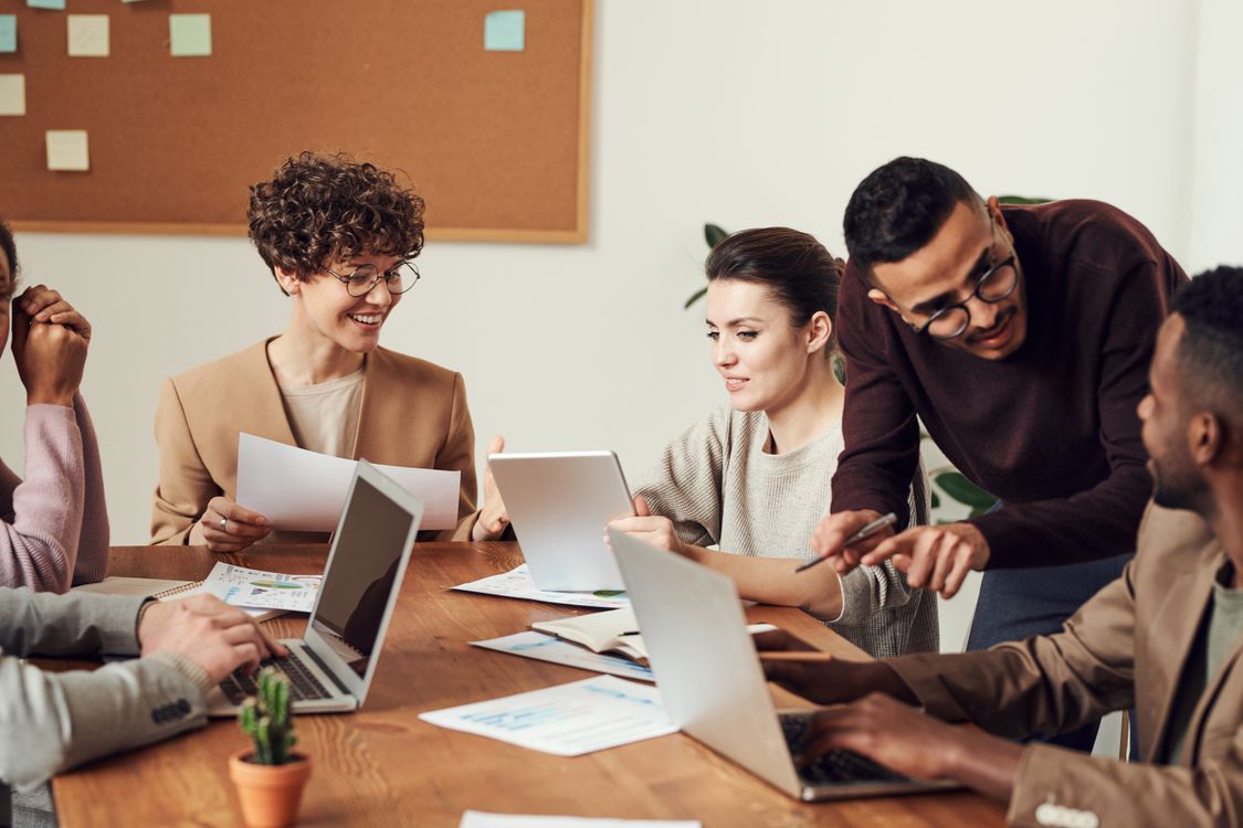 group of people using laptops and tablet around wooden table in office with corkboard
