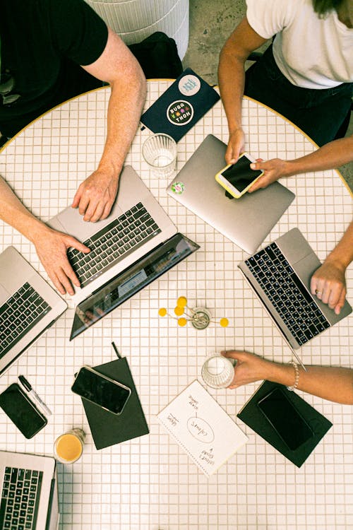 Round table with silver laptops, black smartphones, glass cups, and notebooks on white tiled surface