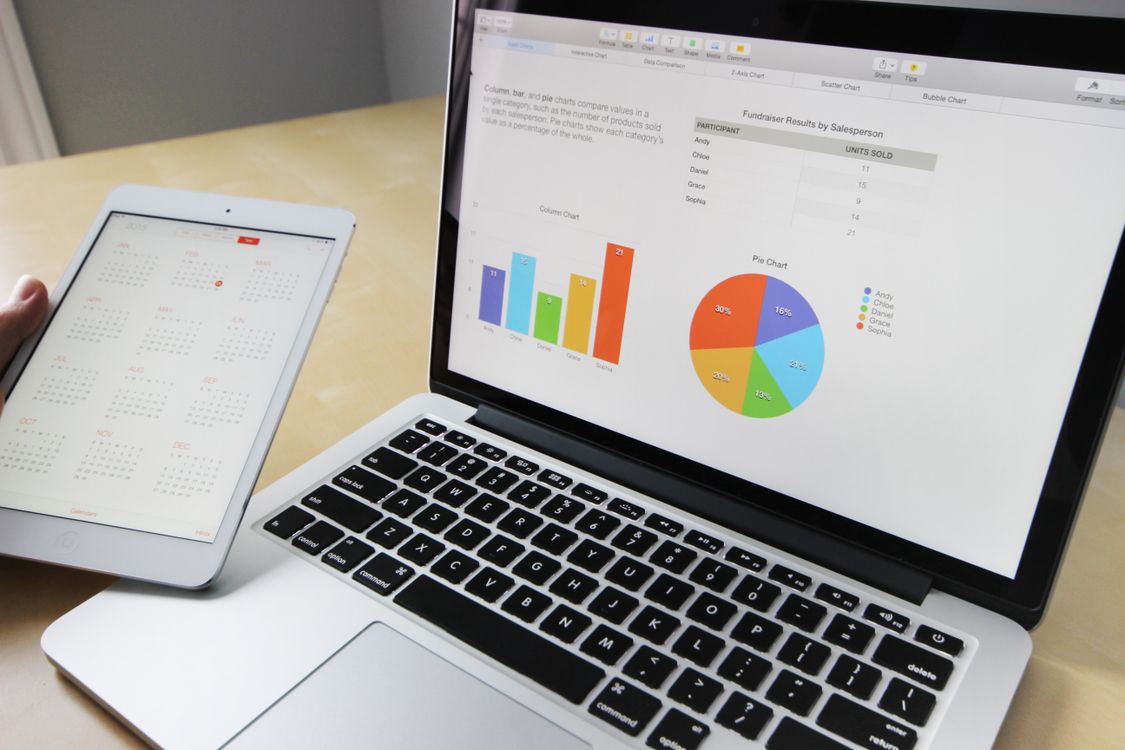 Laptop displaying colorful charts and graphs next to a white tablet showing a calendar on a wooden table