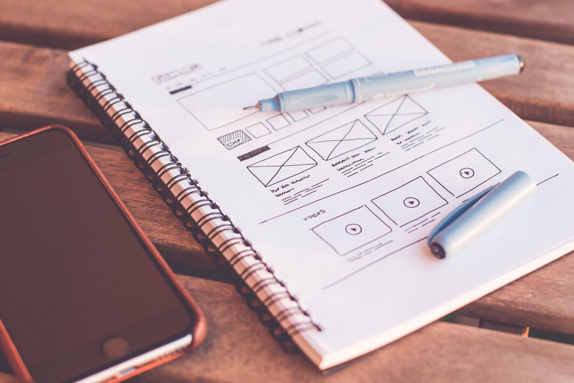 spiral notebook with paper wireframe sketches, blue pen, and black smartphone on wooden surface