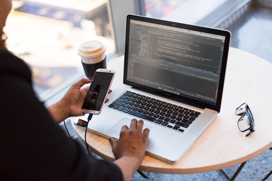 laptop with coding software on screen, smartphone connected by cable, white round wooden table