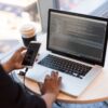 Laptop with coding on screen, person holding smartphone, plastic coffee cup, and black glasses on wooden table