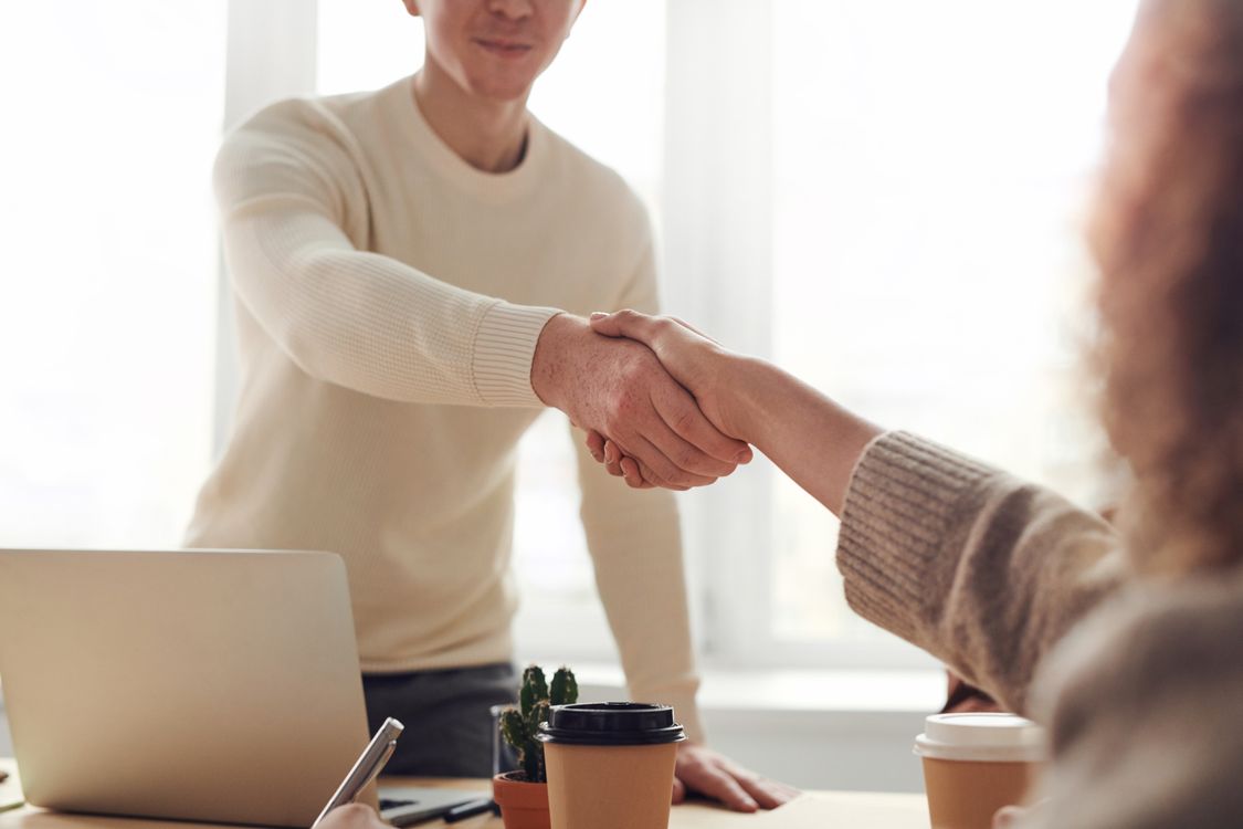 Two people shaking hands over a wooden table with paper coffee cups and a laptop in a bright room