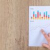 Hand pointing at printed paper with colorful business charts on wooden surface