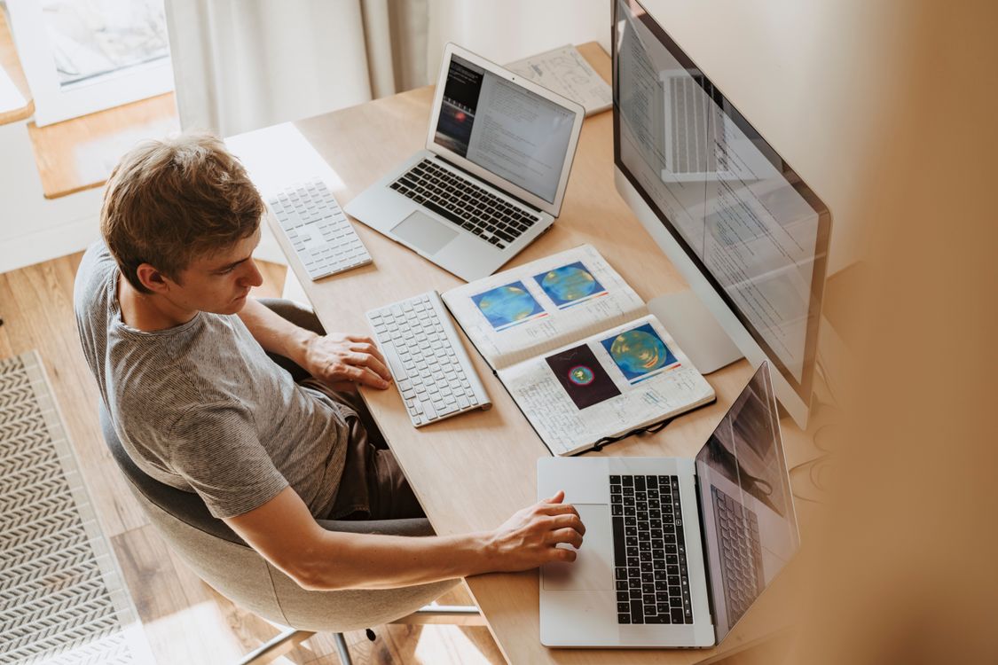 man using multiple silver laptops and a large monitor on a wooden desk with an open notebook