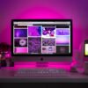 desktop computer with keyboard on white desk with ceramic mug and smartphone holder under pink light
