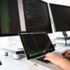 Laptop and dual monitors displaying code on glass and metal screens with hands typing on laptop keyboard
