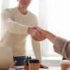 Two people shaking hands, one wearing a beige knit sweater, with paper coffee cups on a wooden table