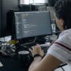 Person typing on black keyboard with black monitor displaying code on glass desk