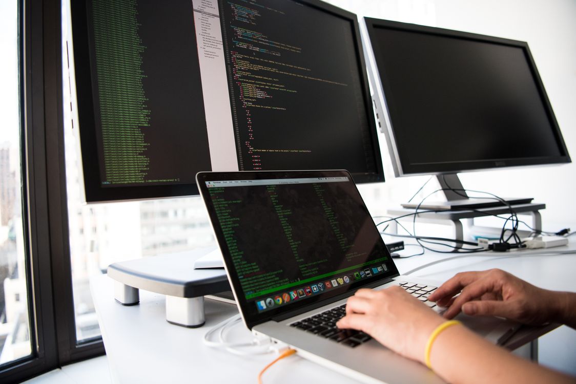 Laptop and dual monitors displaying code on glass screens with metal stands in a bright office