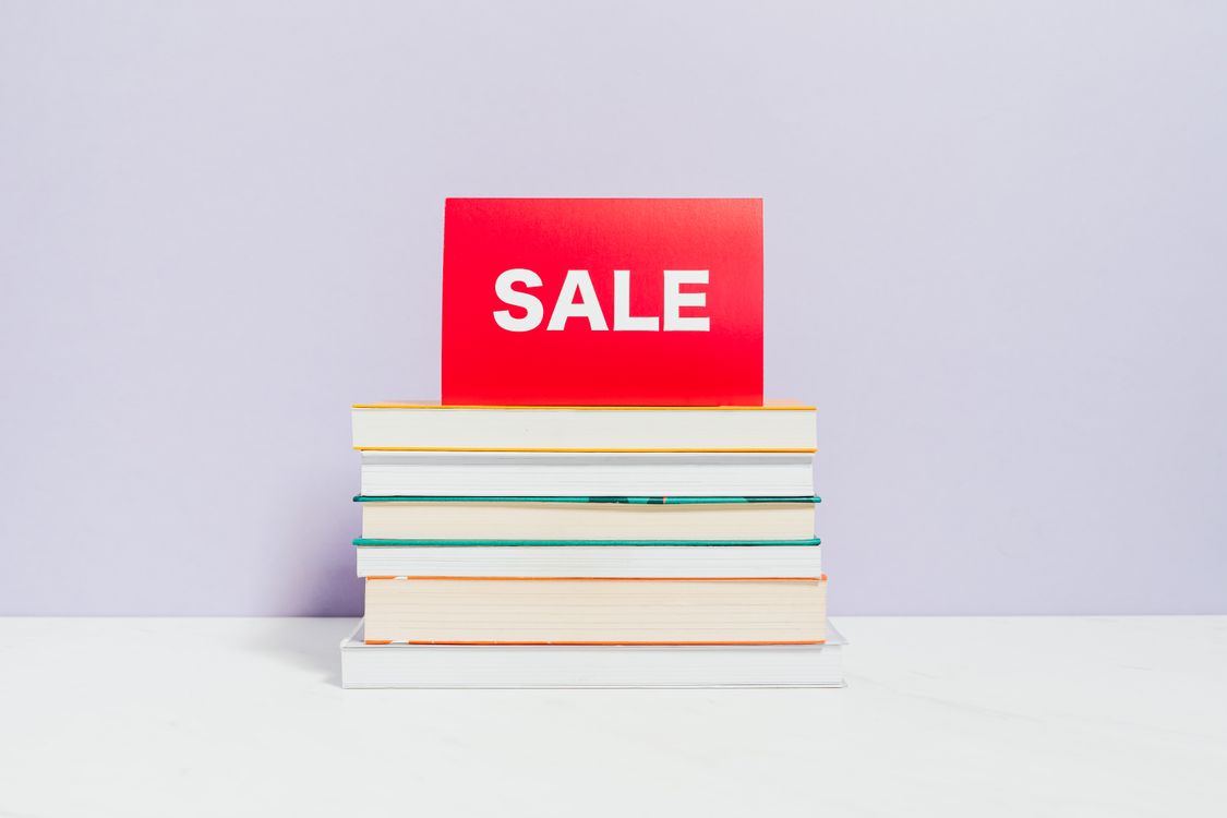 Stack of hardcover books with a red paper sale sign on top on a white surface