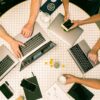 Overhead view of people using laptops and smartphones around a round tiled table with notebooks and glasses, in white and…