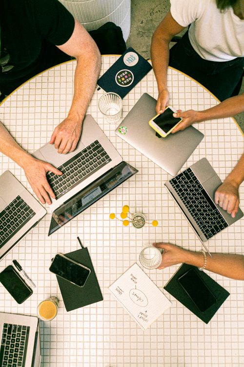 Overhead view of people working on silver laptops and phones around a white tiled table with glasses and a notebook.