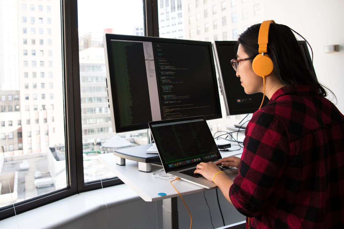 Person using laptop and dual monitors with code, wearing orange headphones and red plaid shirt