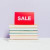 Stack of hardcover books topped with a paper SALE sign, on a white marble surface against a lavender wall, red card